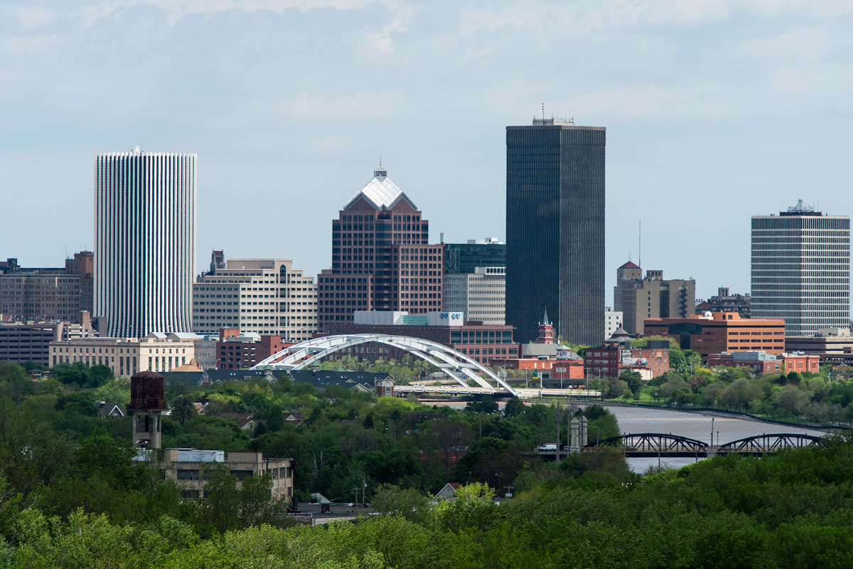 A city skyline featuring a bridge and trees, with the University of Rochester campus in the foreground.