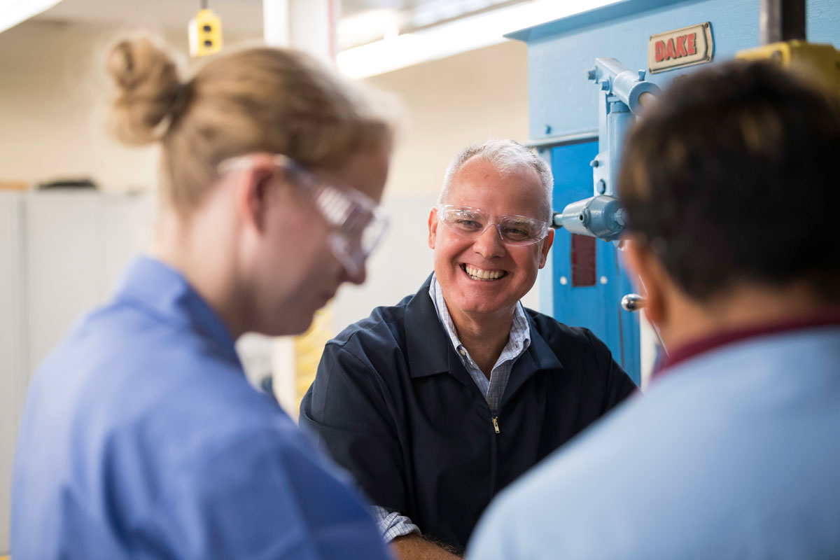 A man engages in conversation with two women in a factory setting, all smiling, showcasing teamwork at the University of Rochester.