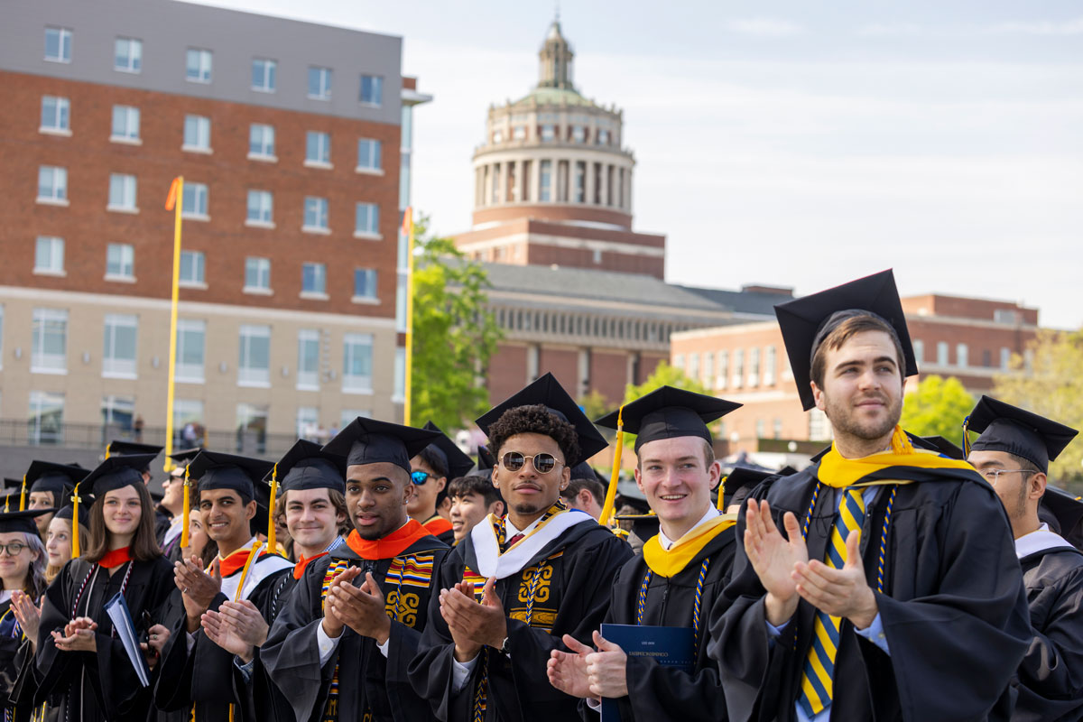 A group of University of Rochester graduates applauding joyfully in front of a historic building during their commencement ceremony.